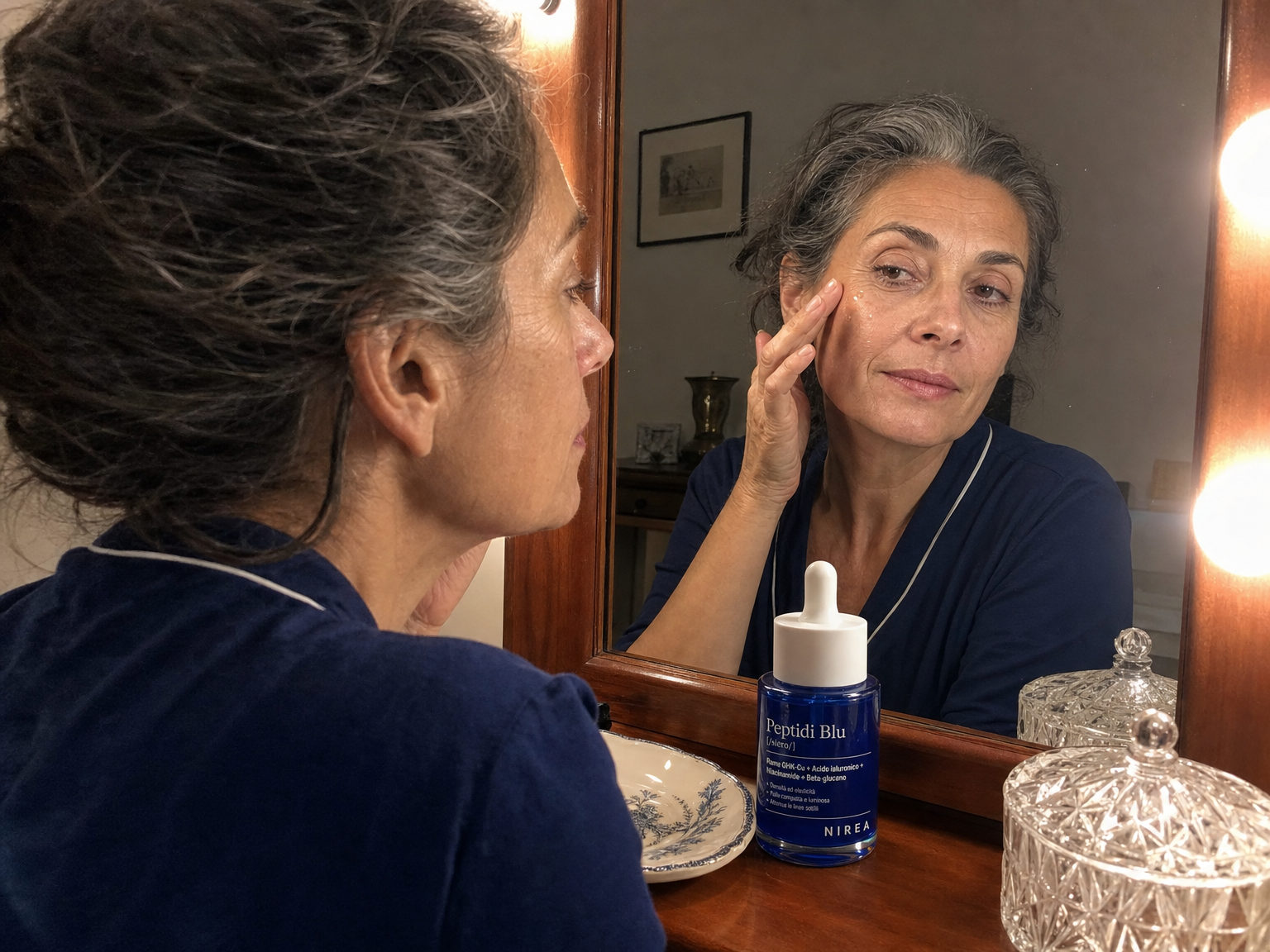 Donna con capelli argento davanti allo specchio del bagno, mentre si applica il siero sul viso con luce calda